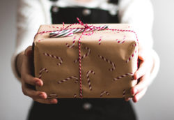 Hands hold a wrapped gift decorated with candy cane patterns and tied with red and white string, set against a blurred neutral background.